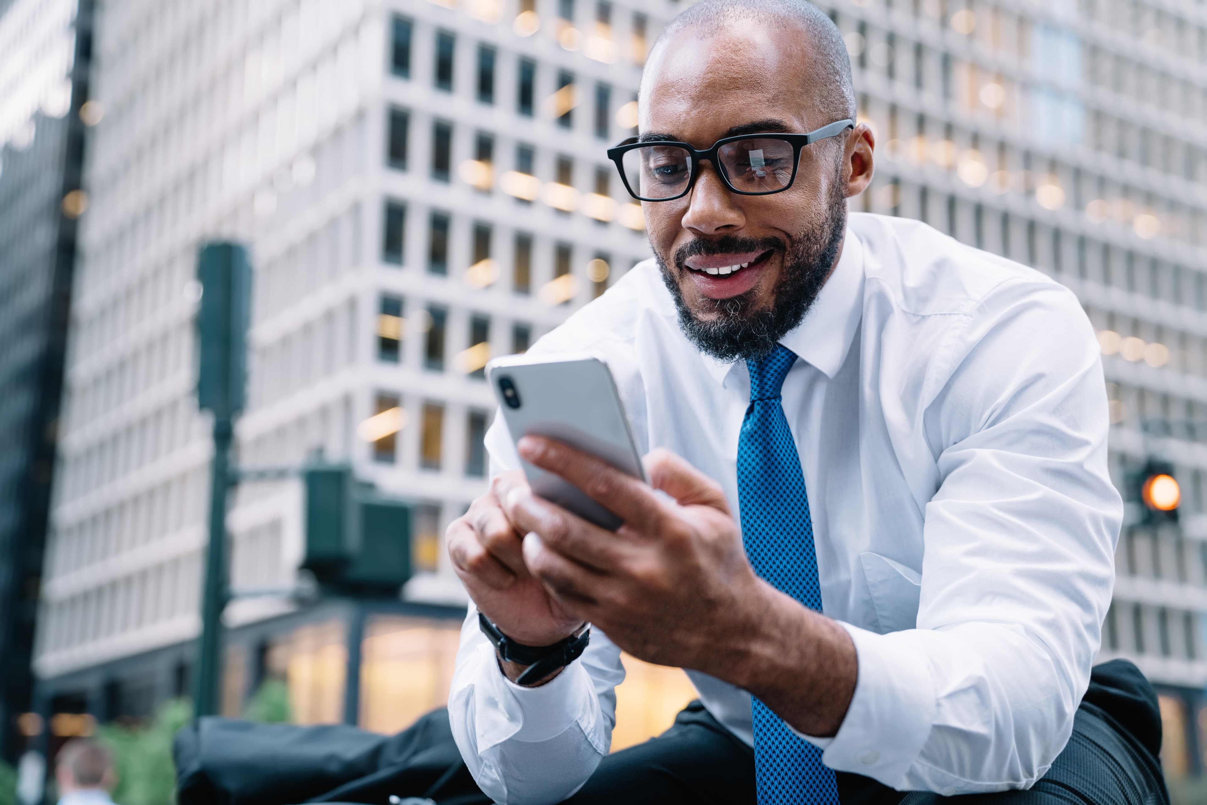 From below cheerful elegant bald African American executive man in white shirt and blue tie browsing mobile while sitting on street stairs on background of blurred business center